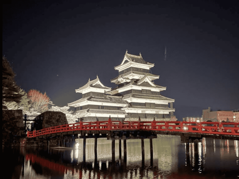 Verlicht Matsumoto-kasteel in Japan met rode brug ernaartoe, gereflecteerd in het water tijdens de nacht.