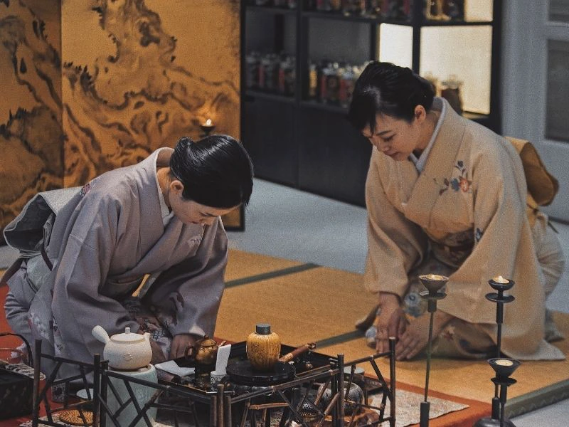 Twee vrouwen in traditionele kimono voeren een Japanse theeceremonie uit, knielend op tatami-matten met theekommen en ceremoniële gebruiksvoorwerpen voor zich.