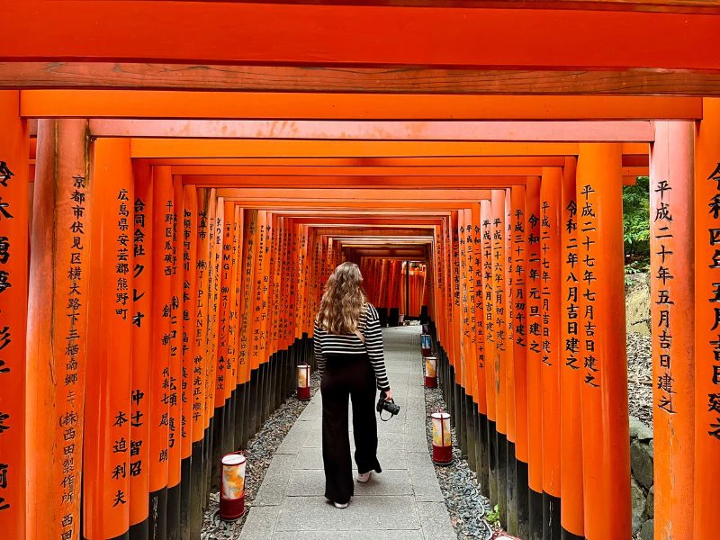Vrouw met lang haar en een gestreepte trui loopt door de iconische rode poortjes van het Fushimi Inari-taisha heiligdom in Kyoto. Ze houdt een camera in haar hand. De poortjes staan dicht op elkaar en zijn bedrukt met zwarte Japanse karakters. De compositie leidt het oog naar het midden van het pad dat tussen de poortjes doorloopt.