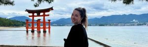 Een reizigster kijkt lachend om in de camera. De rode tempel bij Miyajima eiland steekt uit boven het water. Links daarvan zie je strand met veel groen erachter en rechts in de verte is het bergachtig.