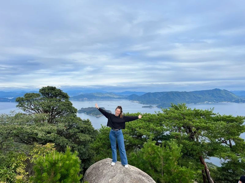 Vrouw op een rots met armen gespreid, op de achtergrond zie je bomen, bergen en water