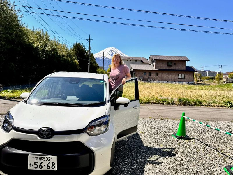 Een reizigster stapt uit een witte Toyota met Japanse kentekenplaat op een zonnige dag, met op de achtergrond Mount Fuji, enkele huizen en groene velden. Rechts naast de auto staat een groene verkeerskegel.