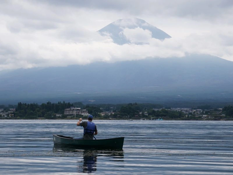 Een persoon in een blauwe reddingsvest peddelt alleen in een groene kano op rustig water. Op de achtergrond is een Japanse stad zichtbaar langs de oever, met daarboven een grotendeels door wolken omhulde Mount Fuji. De sfeer is bewolkt en kalm, met reflecties van de kano en bergen in het water.