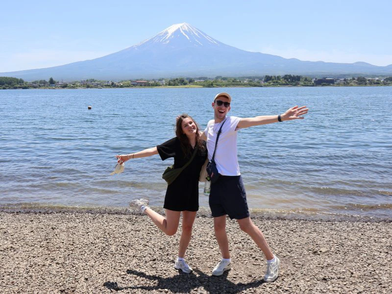 Twee reizigers poseren vrolijk aan de oever van een meer met Mount Fuji op de achtergrond, onder een strakblauwe lucht. De vrouw draagt een zwart jurkje en houdt haar armen gespreid, de man draagt een witte top en donkere shorts. Op de voorgrond ligt een smalle kiezelstrook, op de achtergrond is het helderblauwe water te zien met groene bossen aan de overkant.