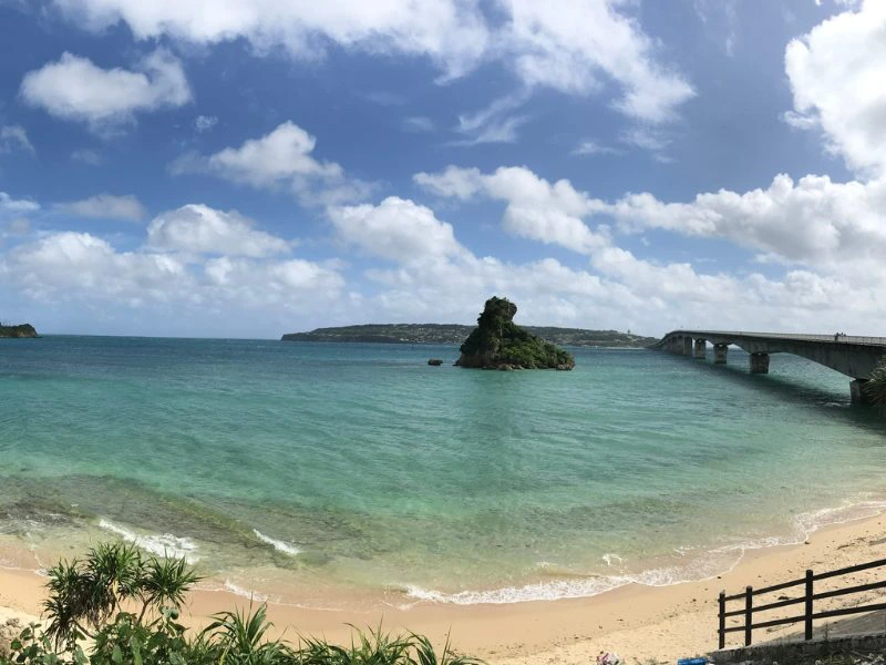 Van bovenaf zie je een geel zandstrand met helder groen/blauw zeewater met in de verte een rots die uitsteekt boven de zee en rechts daarvan een brug over het water. Rechts onderin is een trap te zien. De lucht kleurt blauw met witte wolken.