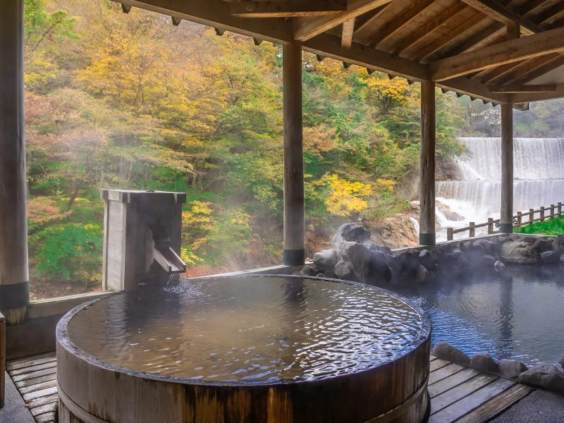 Een rustgevende Japanse onsen met twee warme baden, één rond houten bad op de voorgrond en een groter bad omgeven door natuurlijke stenen aan de rechterkant. De onsen is overdekt met een houten dak en biedt uitzicht op een waterval en herfstkleurige bomen in de achtergrond. Stoom stijgt op uit het warme water, wat de serene en natuurlijke sfeer versterkt.