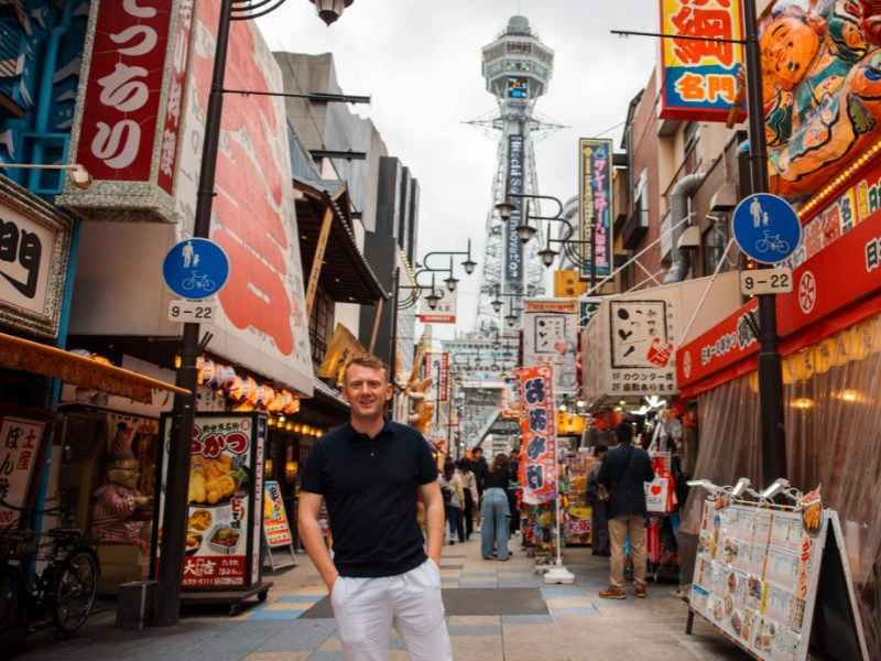 Man poseert lachend met handen in zijn zakken in een kleurrijke winkelstraat in Osaka, Japan. Op de achtergrond staat de Tsutenkaku-toren, omringd door grote Japanse uithangborden en felle neonreclames. Het is overdag en er lopen verschillende mensen door de smalle straat vol restaurants en kraampjes.