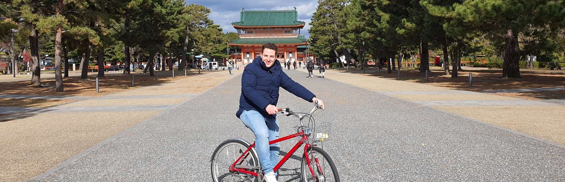 Man fietst op een rode fiets in een brede laan met bomen, richting een traditioneel Japans tempelgebouw in Kyoto.