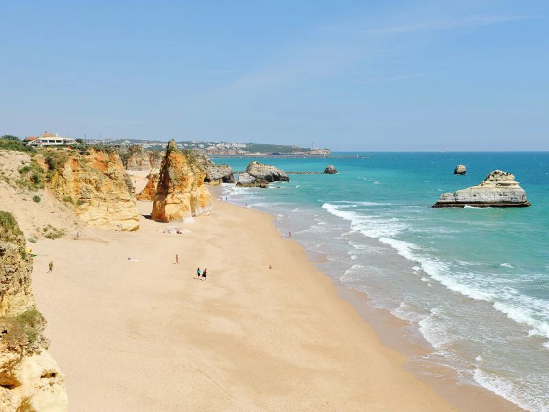 Breed zandstrand met rotspartijen langs de kust en turquoise zee onder een heldere lucht.