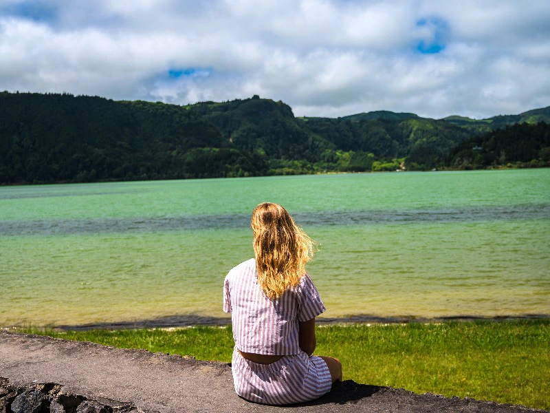 Vrouw zit met de rug naar de camera en kijkt uit over een groen meer met beboste heuvels.