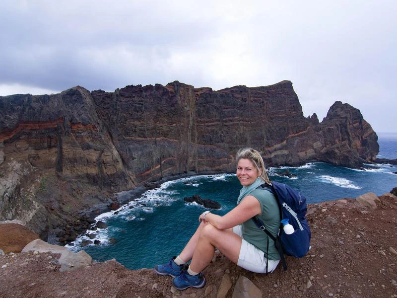 Vrouw met rugzak zit op een rots en kijkt uit over een ruige klifkust met zee.