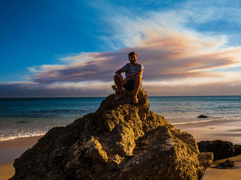 Reiziger zit op een steen op het strand in Portugal met een blauwe lucht op de achtergrond