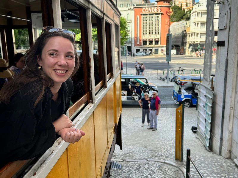 Vrouw leunt lachend uit een geel historisch tramraam in een zonnige stad, met een plein en het gebouw 'Condes' op de achtergrond