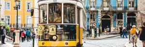 Een klassieke gele tram rijdt door een levendige straat in Lissabon, omringd door kleurrijke gebouwen met tegelfacades en voetgangers die langs de stoep lopen