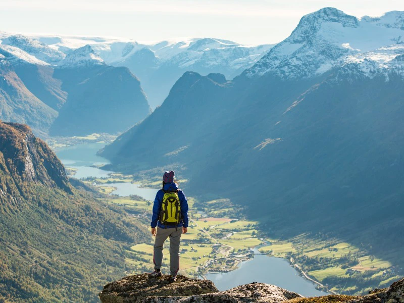 Wandelaar met gele rugzak staat op een rots en kijkt uit over een fjord met besneeuwde bergen.