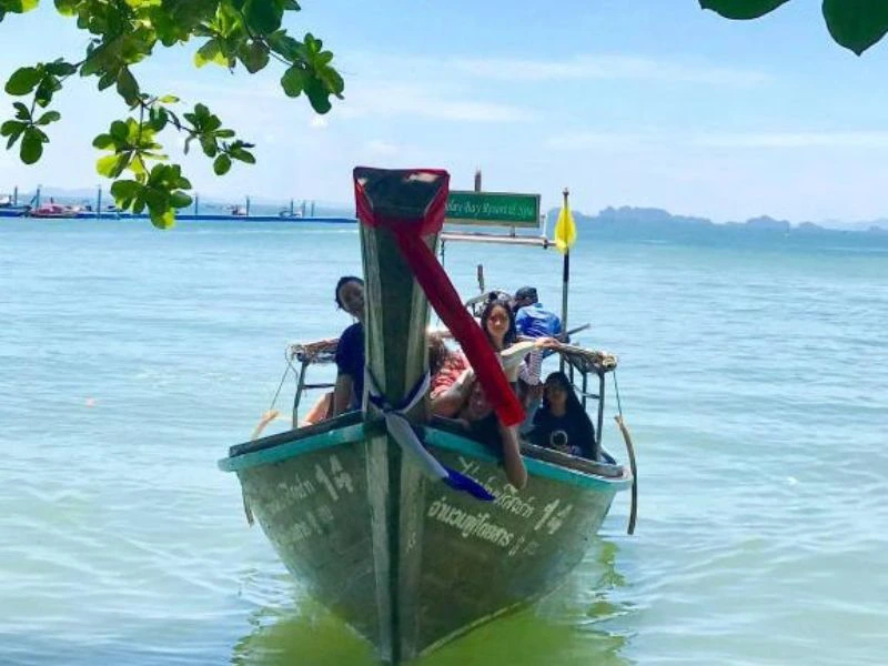 Longtailboot met zes lachende mensen op zee, versierd met linten en vlaggetje. Op de achtergrond kalkstenen eilandjes en andere boten, met takken van een boom bovenin beeld.