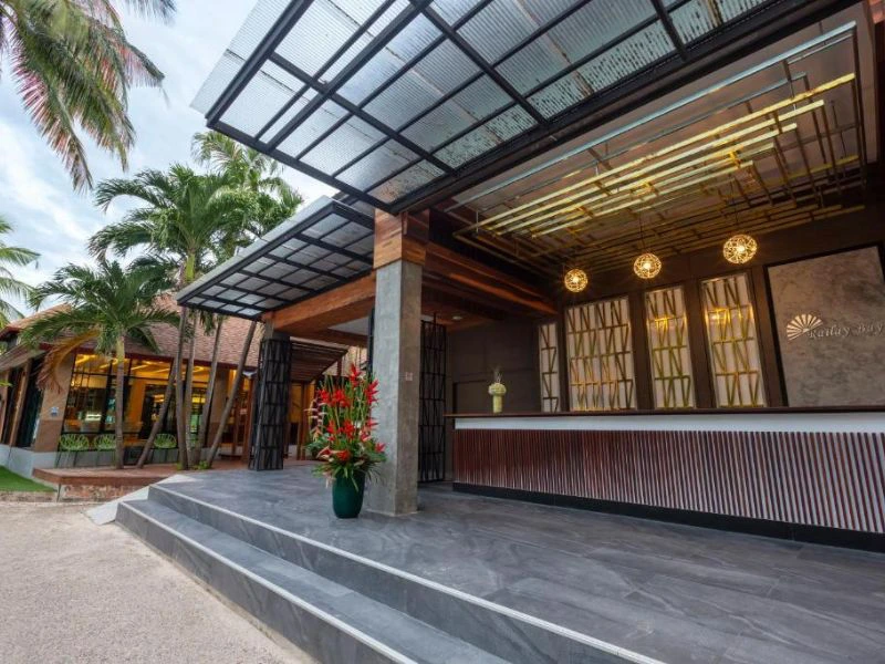 Een moderne hotellobby met een overdekte entree, houten details en sfeervolle verlichting. Voor de ingang staat een grote pot met tropische bloemen en op de achtergrond wuiven palmbomen.