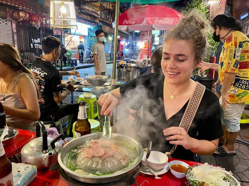 Een vrouw grilt vlees op een hete pot aan een rood tafeltje op een drukke nachtmarkt. Op tafel staan groenten, sausjes, een bierfles en een theepot. Er zitten en lopen mensen om haar heen en op de achtergrond hangen lampjes en staan kleurrijke parasols.