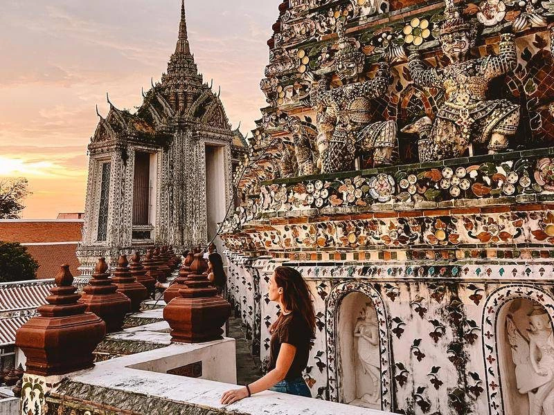 Een vrouw kijkt uit over een rijkversierde tempel in Bangkok met kleurrijke mozaïeken en beelden van wachters. Op de achtergrond is een tweede tempelgebouw te zien met een spitse toren, terwijl de lucht warm kleurt door de ondergaande zon.