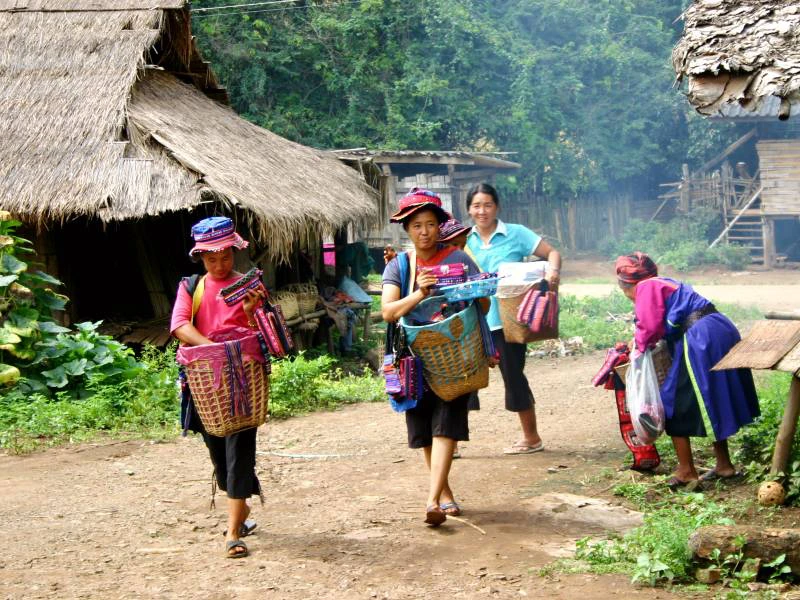 Vrouwen van een bergvolk in Noord-Thailand lopen over een stoffig pad door hun dorp met rieten daken en houten huizen. Ze dragen kleurrijke kleding en hoeden, en hebben manden vol handgemaakte spullen zoals geweven tassen en doeken bij zich. Op de achtergrond zie je meer dorpsbewoners en eenvoudige houten hutten omringd door jungle. Een momentopname van het lokale dorpsleven.