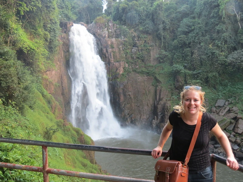 Vrouwelijke reiziger poseert langs een reling met op de achtergrond een hoge waterval, omringd door dichte junglebegroeiing aan beide zijden.