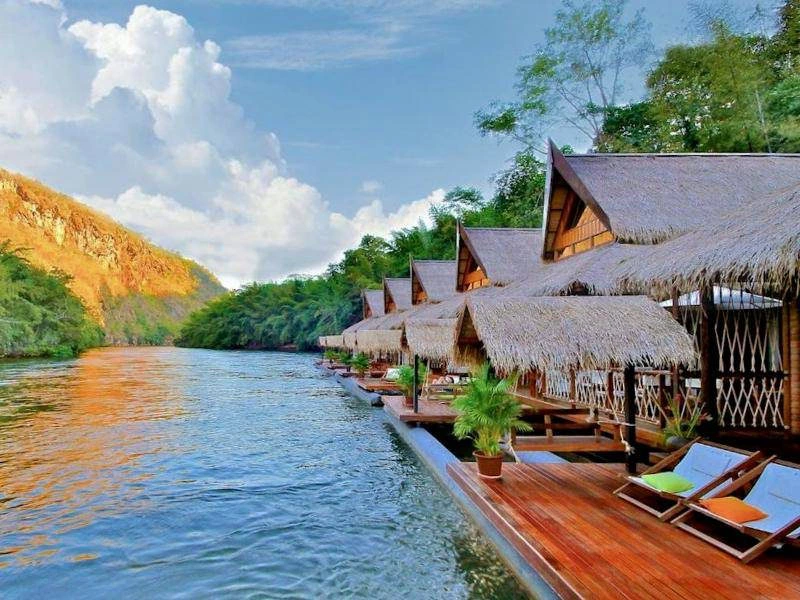 Drijvende houten bungalows met rieten daken aan een rustige rivier, omringd door groene jungle en heuvels. Voor elke bungalow staan ligstoelen en planten, met uitzicht op de rivier die oranje kleurt in de avondzon.