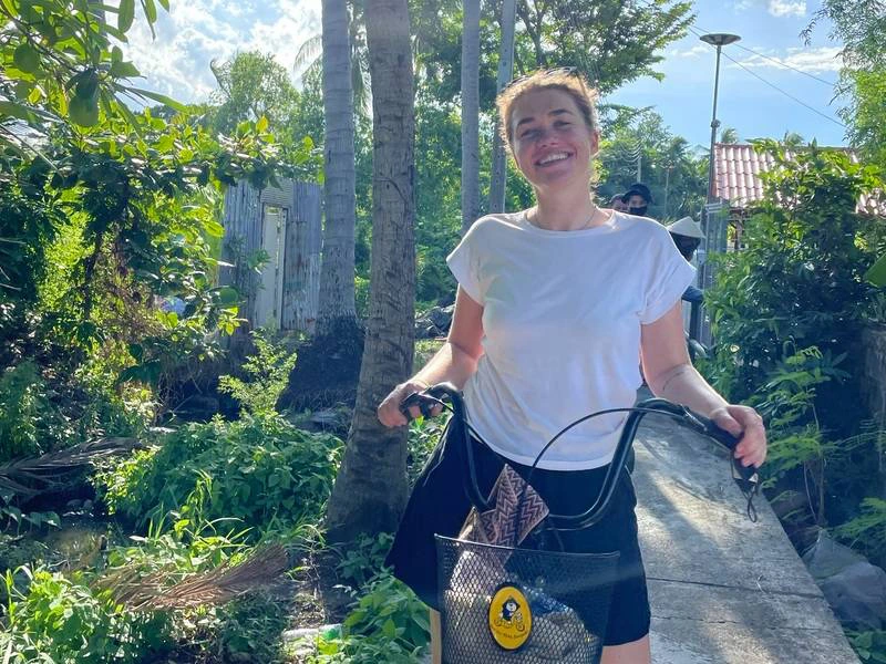 Vrouw met een fiets op een smal pad tussen het groen in een tropische omgeving, met zonlicht dat door de bomen schijnt. Ze lacht en kijkt in de camera.