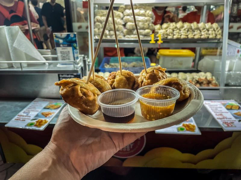 Papieren schotel met gefrituurde dumplings, stokjes en sausjes in iemands hand. Op de achtergrond staat een foodtruck