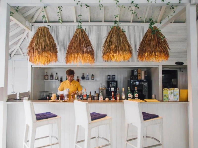 Kleurrijke strandbar met witte inrichting, vier rieten lampen en een barman in een geel shirt die drankjes klaarmaakt achter de bar. Op de bar staan flessen drank en op de barkrukken liggen paarse kussens.