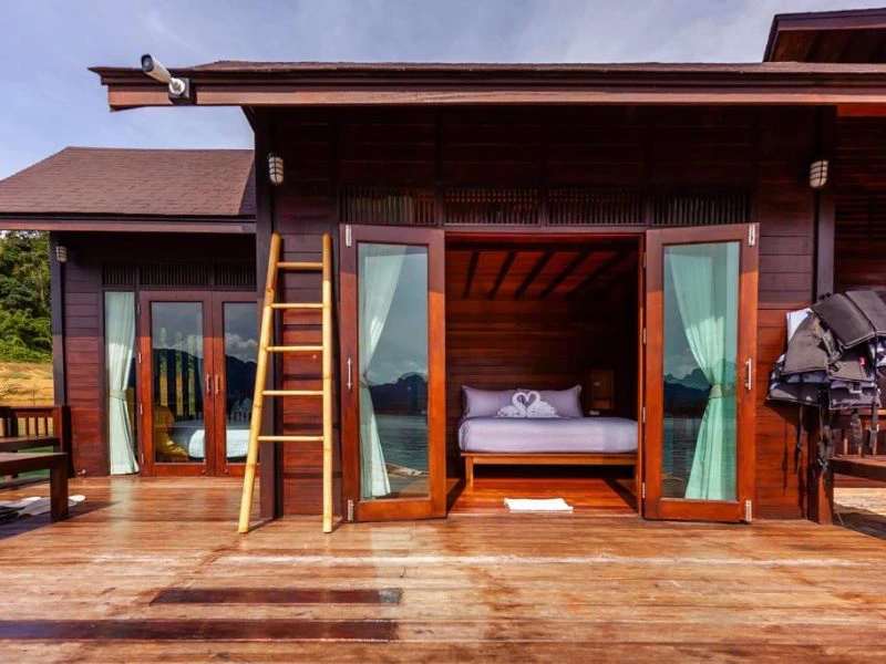 Houten bungalow op het water met openstaande deuren, uitzicht op het meer en bergen op de achtergrond. Binnen staat een bed met handdoekzwanen, buiten ligt een houten vlonder met een trapje en zwemvesten.