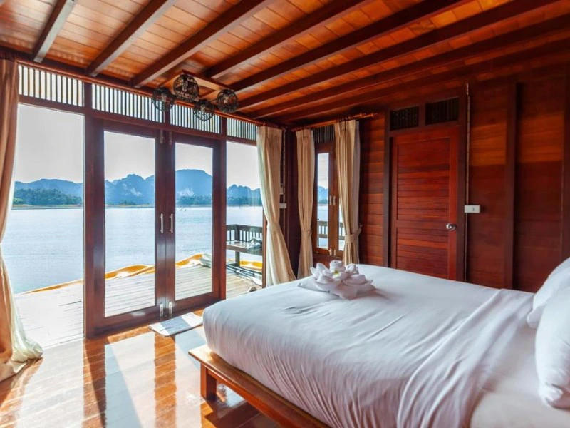 Houten drijvende bungalow met dubbele glazen deuren en lichtgele gordijnen, een bed met opgevouwen handdoeken in zwanenvorm, houten terras aan het water en op de achtergrond bergen en bos.