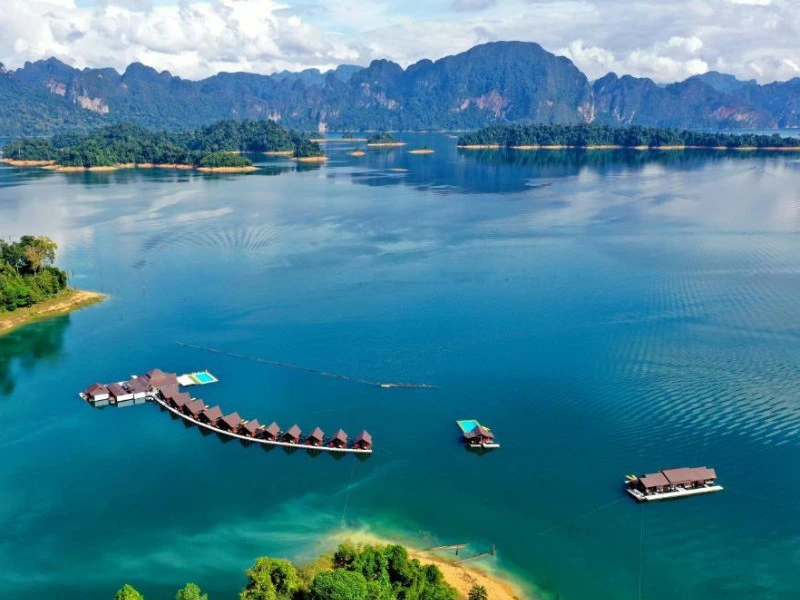 Luchtfoto van een meer met een rij drijvende bungalows in een boogvorm, omringd door helderblauw water, groene eilanden en op de achtergrond een bergachtig landschap met dichte jungle.