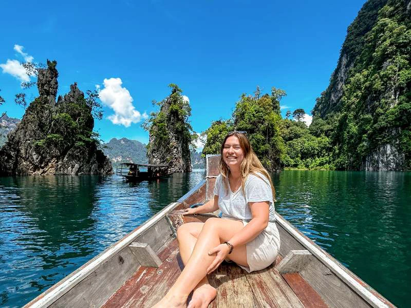 Vrouw zittend op de houten punt van een boot op helder water, omringd door groene kalkstenen rotsformaties en een blauwe lucht met wolken.