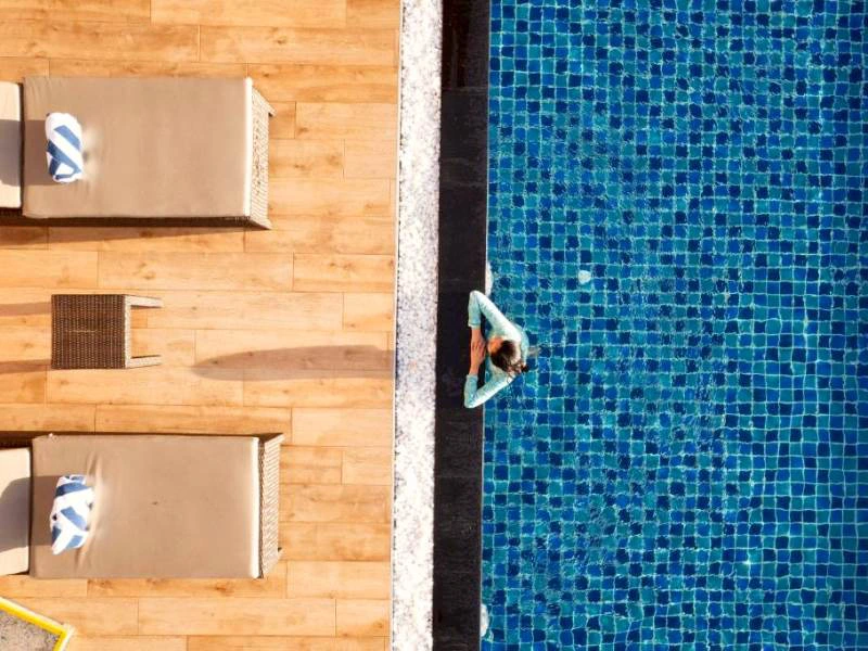 Van bovenaf gefotografeerd: een vrouw hangt ontspannen aan de rand van een strak blauw mozaïektegel zwembad. Naast het zwembad liggen houten ligbedden met opgerolde handdoeken op een houten vlonder.