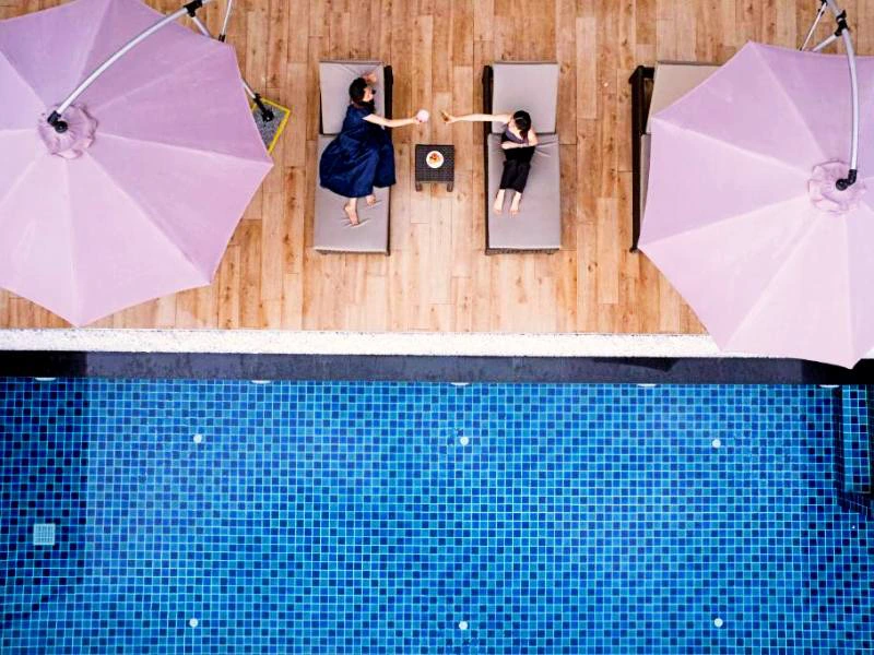 Van bovenaf gefotografeerd: twee personen liggen ontspannen op ligbedden onder roze parasols aan een zwembad met blauwe mozaïektegels. Ze reiken naar elkaar met hun handen, met een tafeltje tussen hen in.