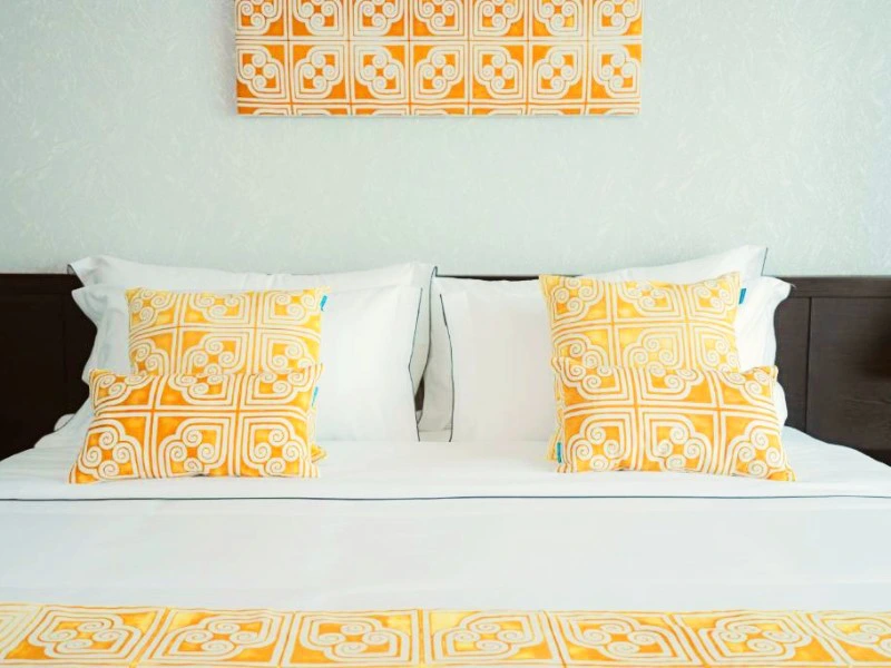 Licht en fris hotelbed met witte lakens, twee geel-wit patroon sierkussens en een bijpassende loper. Boven het bed hangt een kunstwerk in dezelfde stijl als de kussens.
