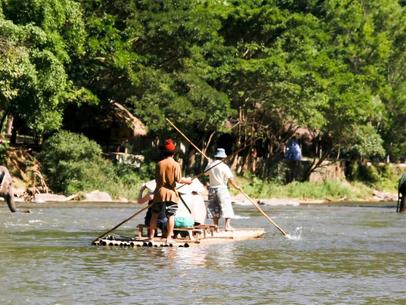 Vier mensen staan op een vlot van bamboe op een rivier, omgeven door groene bomen. Twee van hen gebruiken stokken om het vlot voort te duwen. Op de achtergrond zijn hutten en bomen zichtbaar.