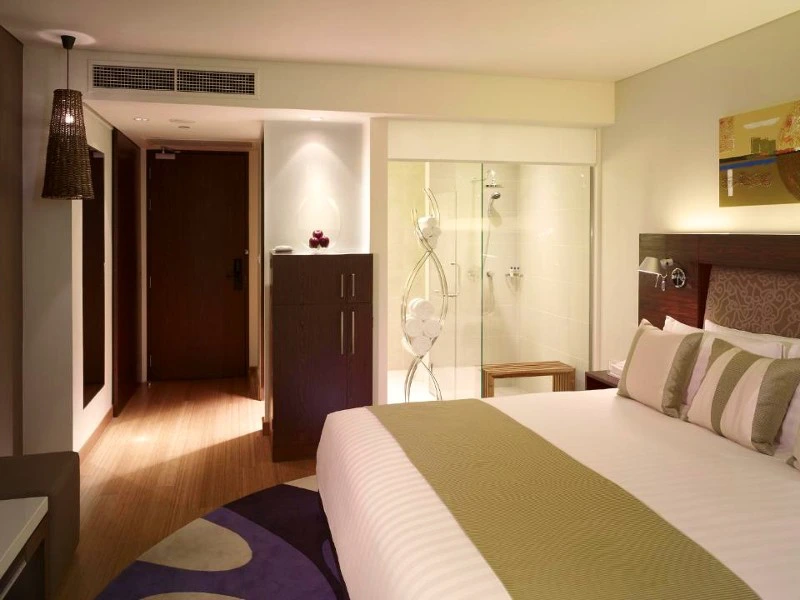 Modern hotelkamer met een groot tweepersoonsbed, kussens en een beige-groene sprei. Rechts achterin is een glazen wand met daarachter een inloopdouche zichtbaar.