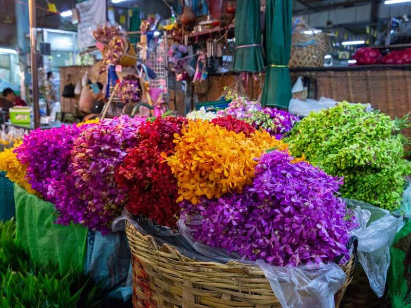 Mand vol kleurrijke bloemen in paars, rood, oranje en groen op een lokale markt, met op de achtergrond stalletjes en handgemaakte spullen.