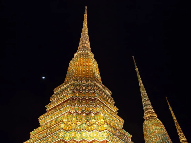 Verlichte stoepa’s van een tempelcomplex in Thailand, ’s avonds gefotografeerd tegen een donkere hemel met een maan in de achtergrond.