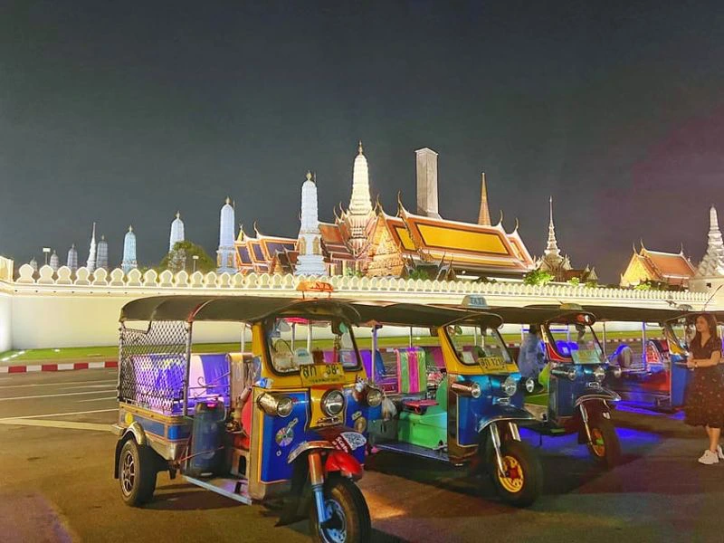 Twee reizigers zitten lachend achterin een tuk-tuk terwijl ze 's avonds door de levendige straten van Bangkok rijden. Om hen heen lichten de neonkleuren op, en op de achtergrond zie je een billboard en het drukke stadsleven.