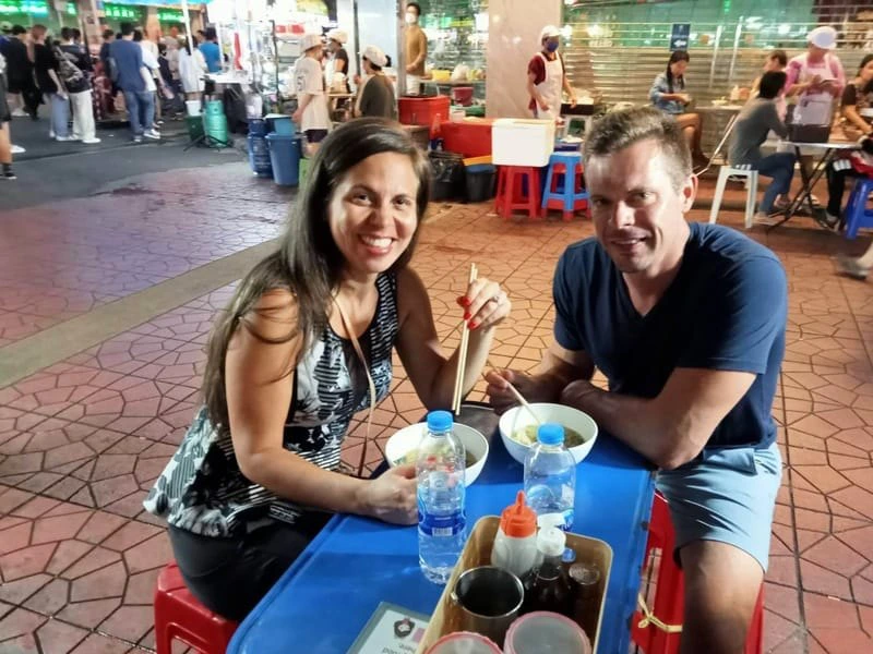 Twee reizigers genieten van een maaltijd aan een klein blauw plastic tafeltje op een levendige avondmarkt in Thailand. Ze lachen naar de camera terwijl ze met stokjes eten; om hen heen zie je locals, andere reizigers en kraampjes met streetfood.