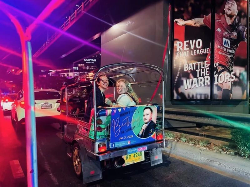 Twee lachende mensen in een kleurrijke tuktuk in het avondverkeer van Bangkok, met neonlichten, een reclamedoek met sporter op de achtergrond en een verjaardagsbanner achterop de tuktuk.