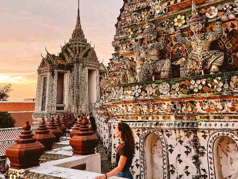 Een vrouw kijkt uit over een rijkversierde tempel in Bangkok met kleurrijke mozaïeken en beelden van wachters. Op de achtergrond is een tweede tempelgebouw te zien met een spitse toren, terwijl de lucht warm kleurt door de ondergaande zon.