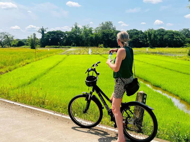 Een vrouw zit op een zwarte fiets op een betonnen weg, met felgroene rijstvelden op de achtergrond. Ze draagt een donkere top, een korte broek met panterprint en een zonnebril. Het is zonnig weer.