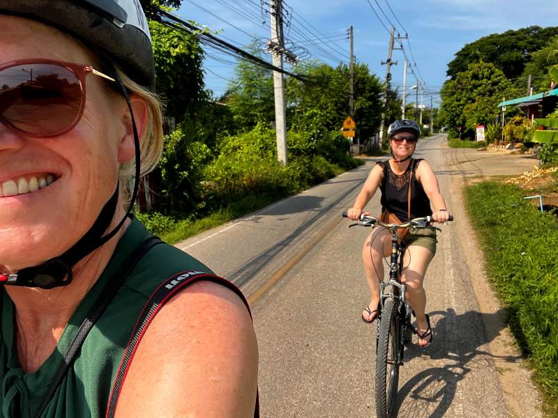 Twee vrouwen fietsen over een rustige weg met helmen op, omringd door groen en elektriciteitspalen.