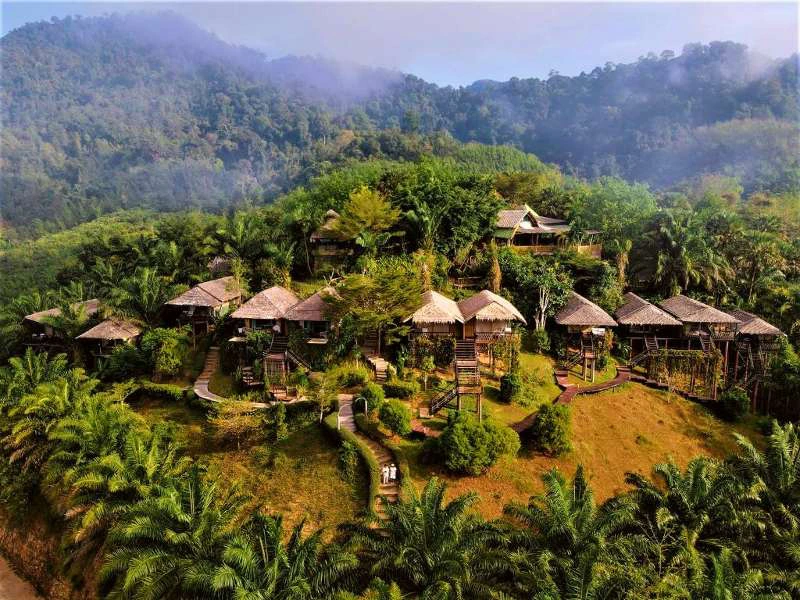 Kleinschalige houten bungalows met rieten daken liggen verspreid op een groene heuvel, omringd door palmbomen en jungle. De huisjes zijn met elkaar verbonden via slingerende paadjes. Op de achtergrond zie je mistige heuvels en dichte bebossing, diep in het Thaise binnenland.
