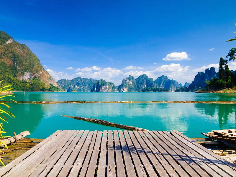 Een houten vlonder aan een turquoise meer, omringd door groene kalkstenen rotsen en een blauwe lucht met enkele wolken.