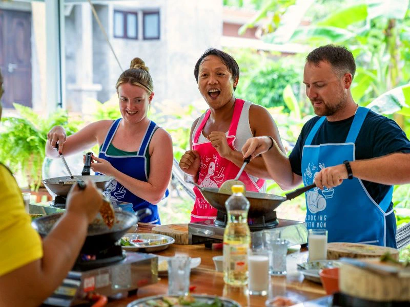 Drie reizigers staan zij aan zij achter een kookstation tijdens een Thaise kookworkshop. Ze dragen kleurrijke schorten en roeren in hun pannen, aangemoedigd door de lokale kok die met een grote glimlach uitleg geeft. Op de achtergrond tropisch groen en een open ruimte die zorgt voor een ontspannen sfeer. Een tafel vol ingrediënten, glazen en flessen olie geeft een levendig beeld van het koken.