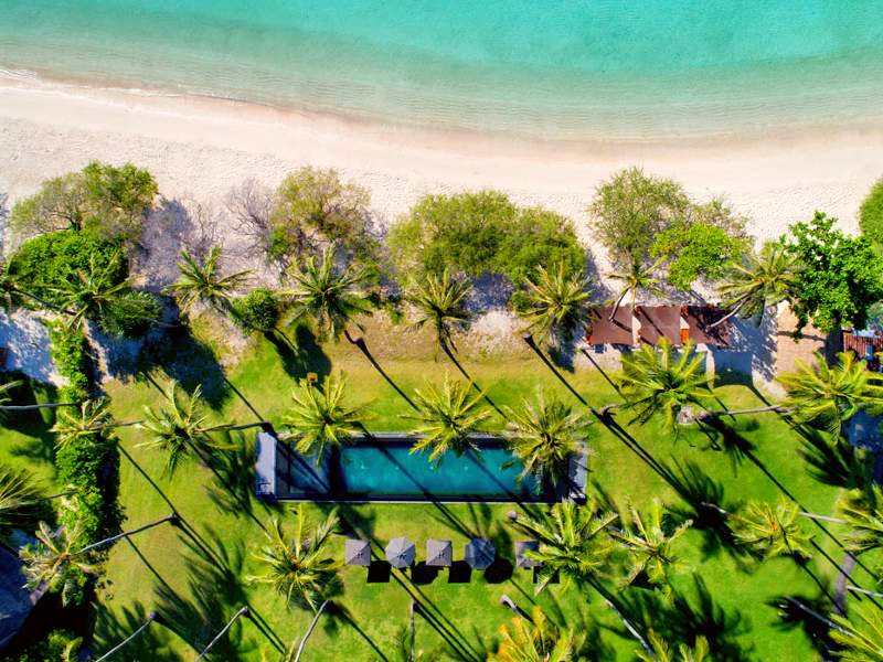 Luchtbeeld recht naar beneden van een strand met blauwe zee, grasvelden met bomen, rieten hutjes en een zwembad.